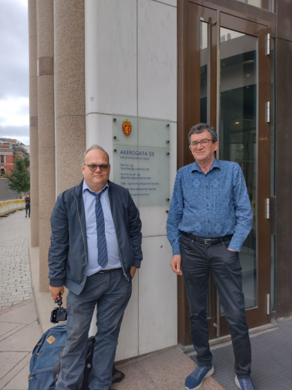 To personer står foran en bygning med et skilt som viser adressen "Akersgata 59" i Oslo.