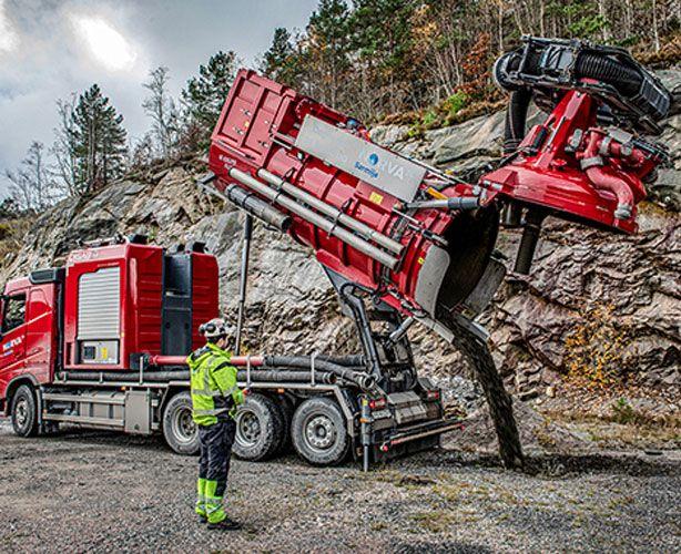 En rød lastebil med en stor betongpumpe som tømmer betong på en steinete byggeplass.