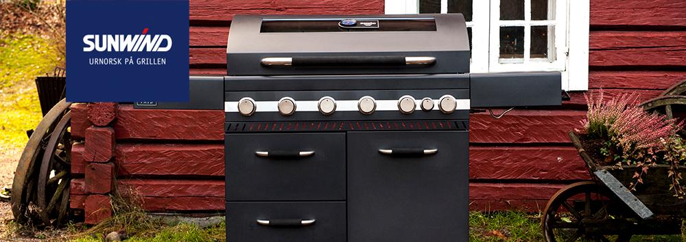 En moderne gassgrill fra Sunwind plassert foran en rød hytte med vinduer.