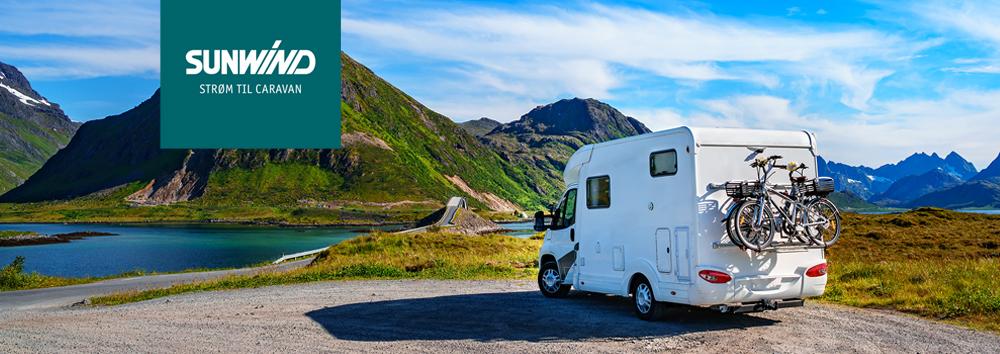 En campingvogn parkert ved en vakker innsjø med fjell i bakgrunnen, med sykler festet bakpå.