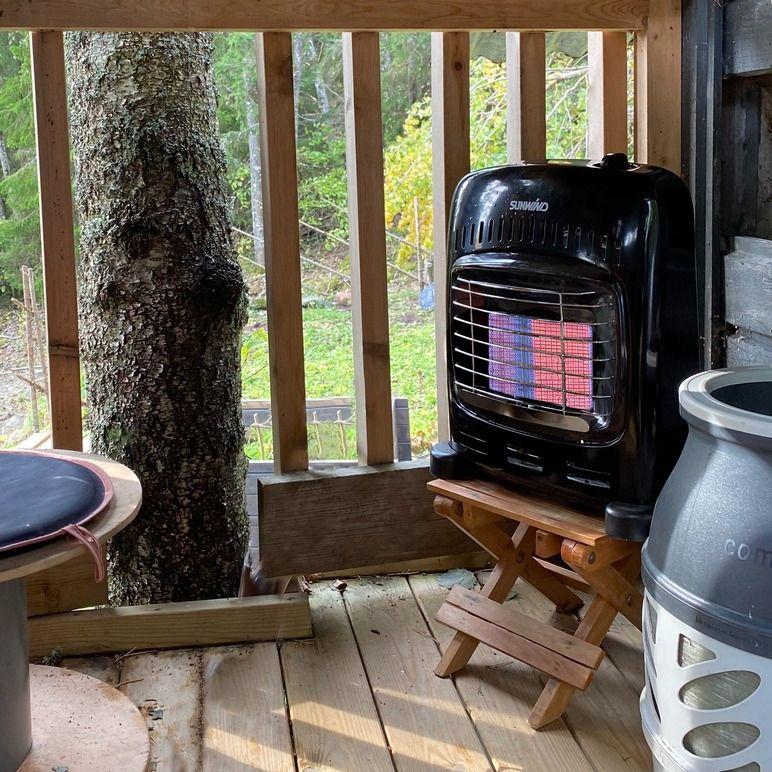 En svart gassovn plassert på en trebenk i en hytte med utsikt mot en grønn hage.