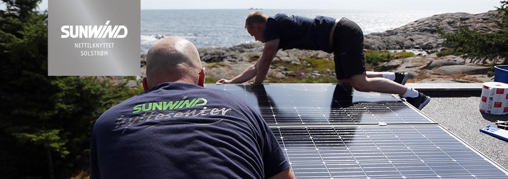 To personer installerer solcellepaneler på et tak med havet i bakgrunnen.