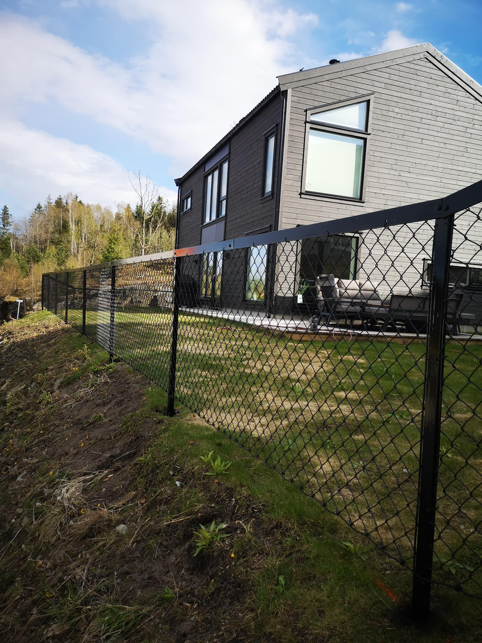 Moderne hus med store vinduer og terrasse, omgitt av et svart gjerde i en grønn hage.