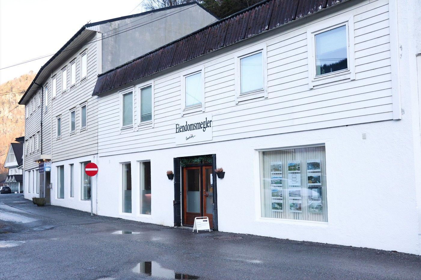 En hvit bygning med flere vinduer og en inngangsdør, beliggende langs en vei i en liten by.