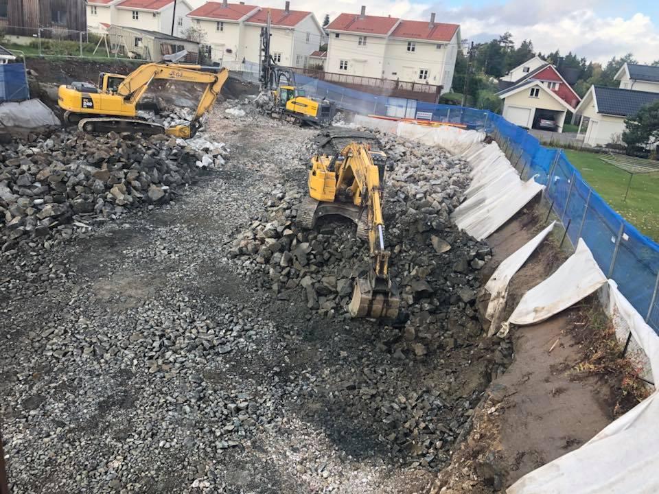 Byggeplass med gravemaskiner som fjerner stein og grus, omgitt av gjerder og hus i bakgrunnen.