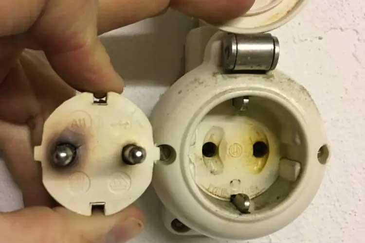 En hånd som holder en elektrisk plugg ved siden av en stikkontakt med synlige brennmerker.