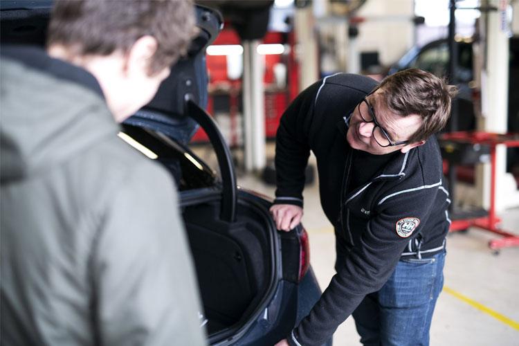 To personer i et verksted, en mekaniker som viser noe i bagasjerommet på en bil.