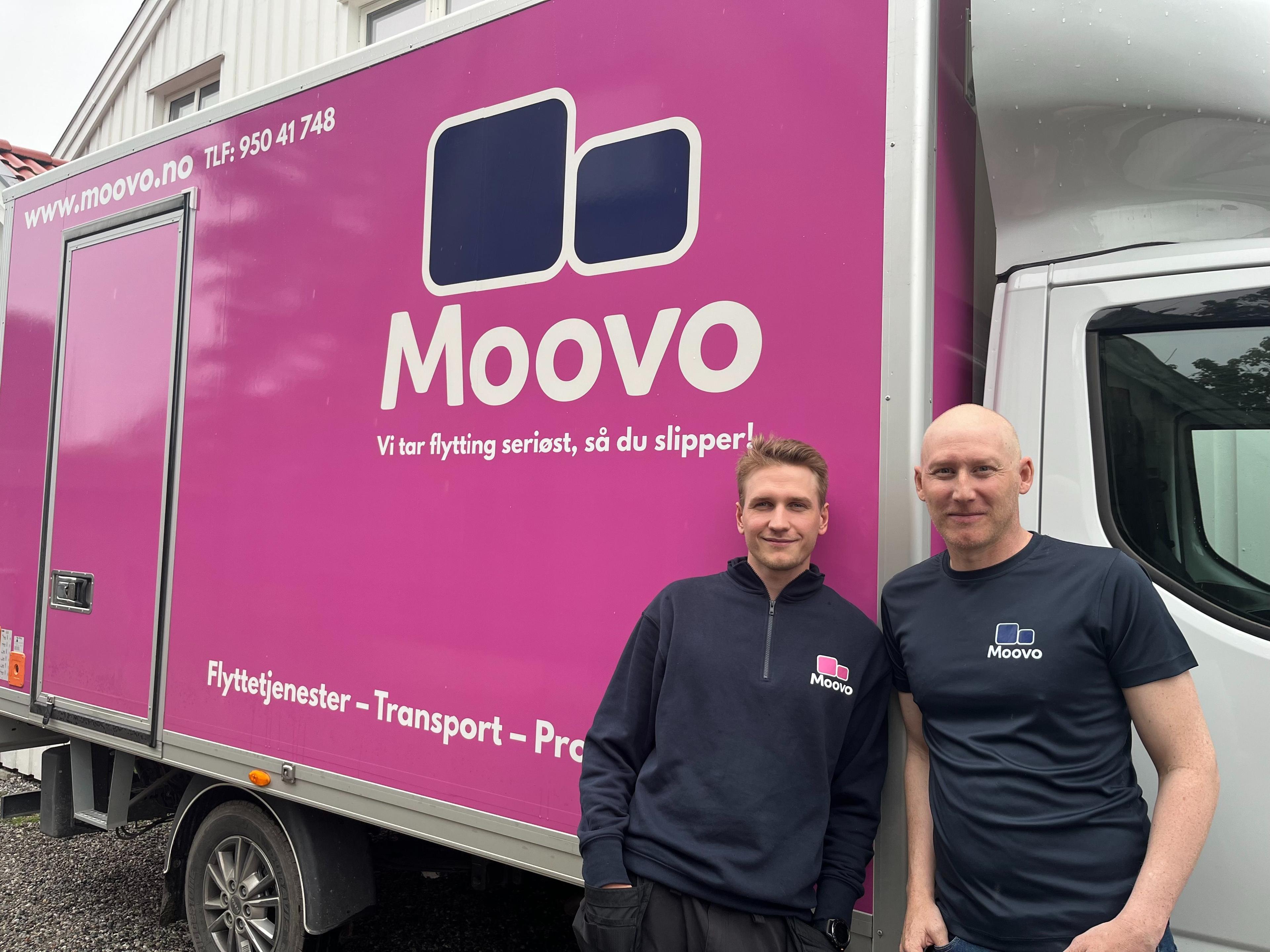 To personer står foran en rosa flyttebil med Moovo-logoen, som tilbyr flyttetjenester og transport.