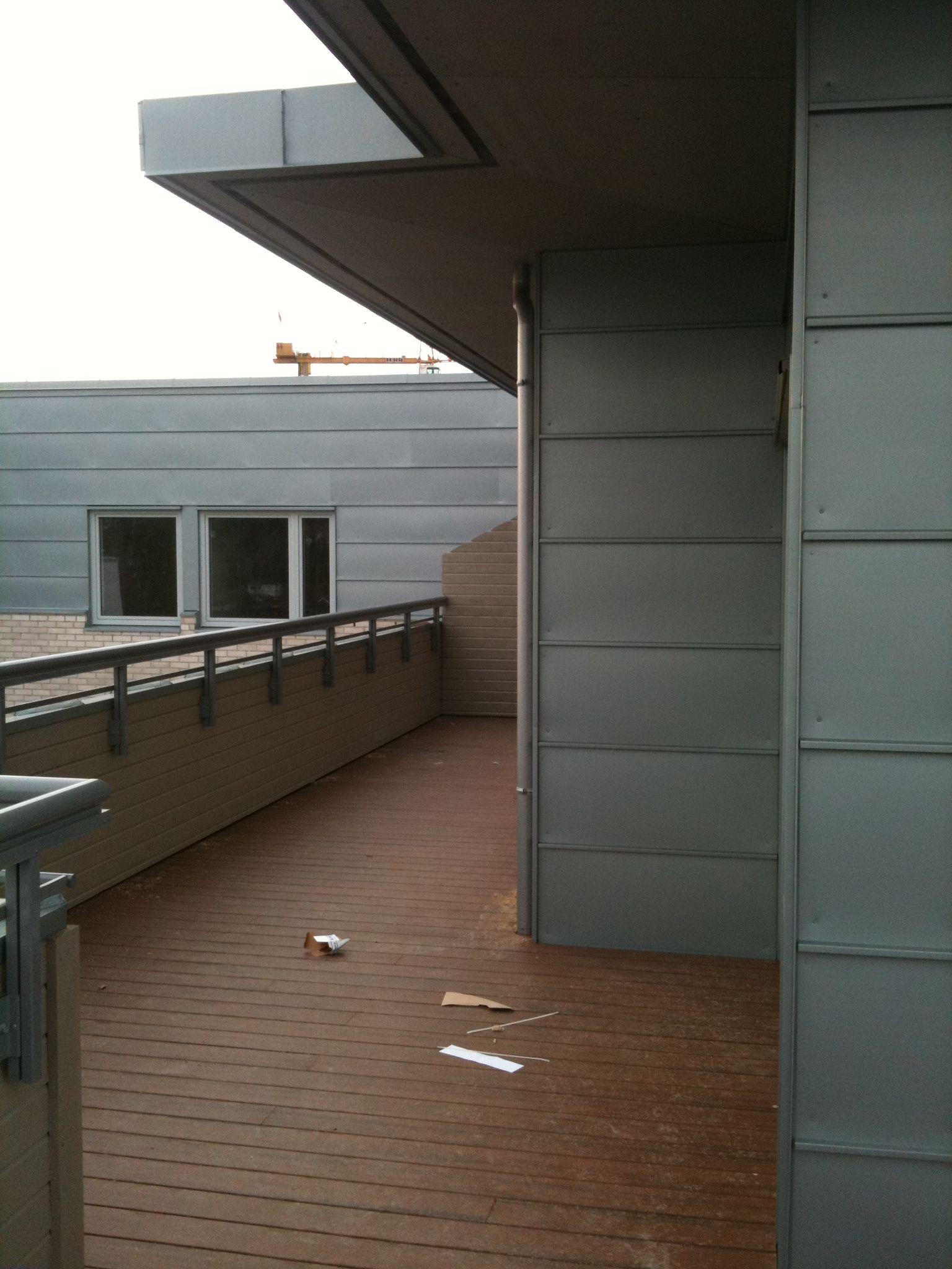 Balkong med tredekk og moderne bygning i bakgrunnen, med grå vegger og vinduer.