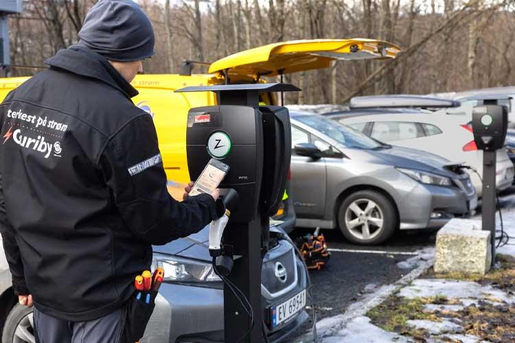En mann bruker en mobiltelefon ved en elbil-lader i en parkeringsplass med flere biler.