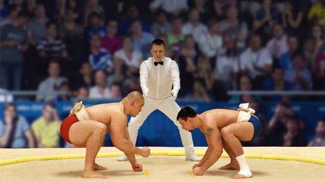 To sumo wrestlers i ringen, med en dommer i hvit dress som overvåker kampen foran et publikum.