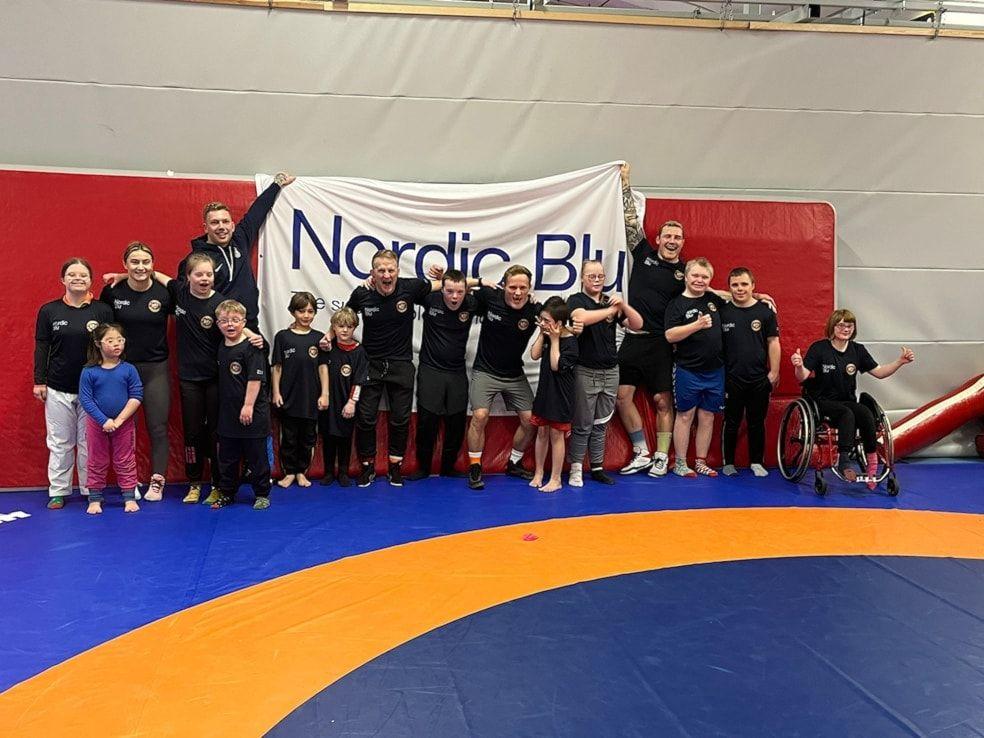 En gruppe unge og voksne brytere poserer sammen med et banner for Nordic Blå i en brytesal.