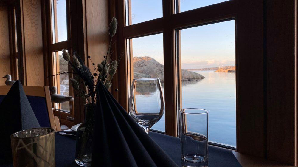Utsikt fra et restaurantbord med glass og servietter, mot en rolig sjø og fjell i bakgrunnen.