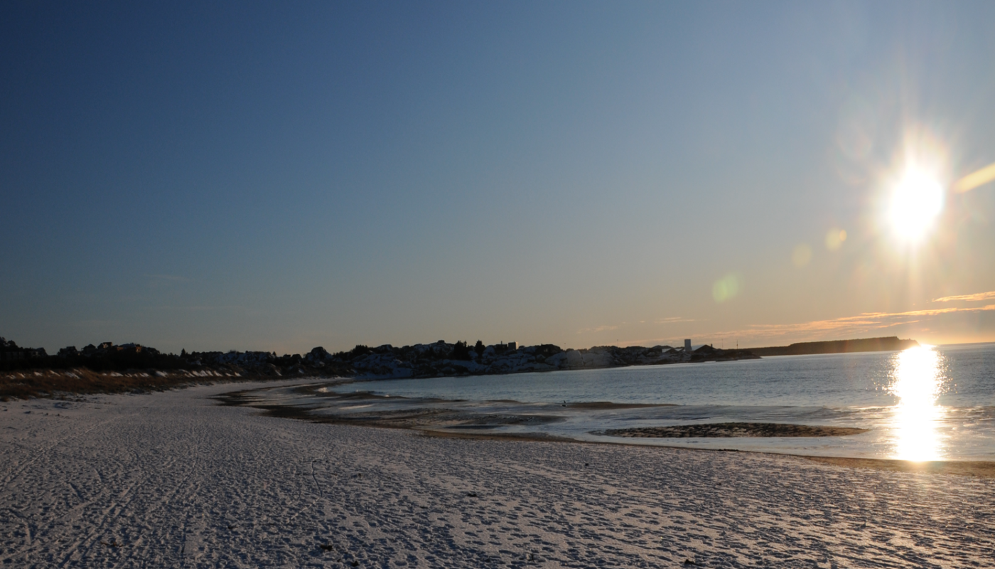 En solfylt strand med snø og hav, med en klar himmel og en by i bakgrunnen.