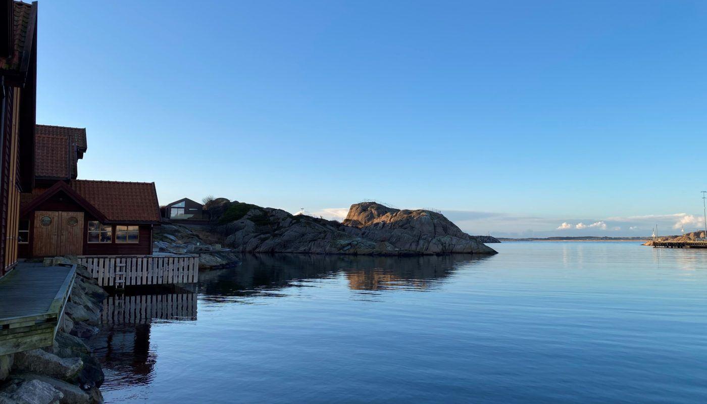 En rolig havn med speilbilde av fjell og bygninger i sollys, omkranset av klart blå himmel.