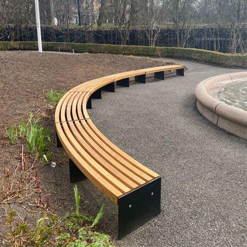 Bueformet benk i tre med svart metallramme, plassert ved en fontene i en park.