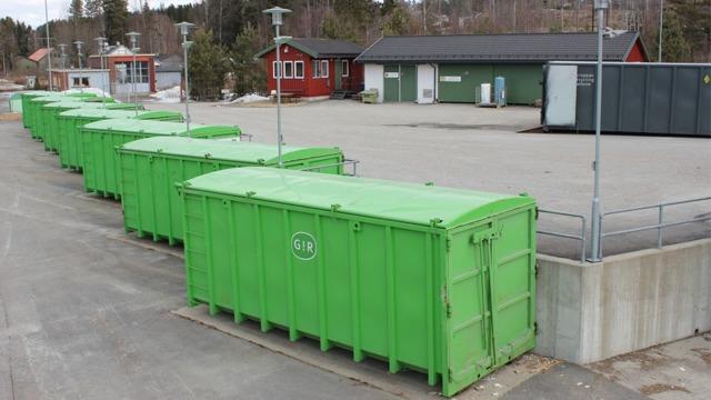 Grønne avfallscontainere på en parkeringsplass med bygninger i bakgrunnen.