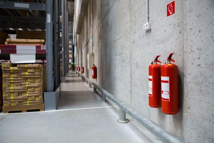 Brannslukkere på veggen i et lager med betongvegg og paller med varer i bakgrunnen.