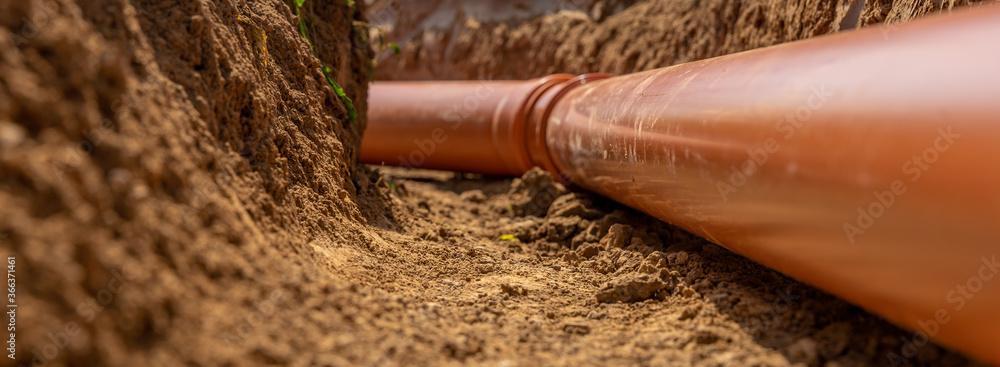 Rørledning i en gravd grøft, dekket av jord og grus, med fokus på den oransje plastikkrøret.