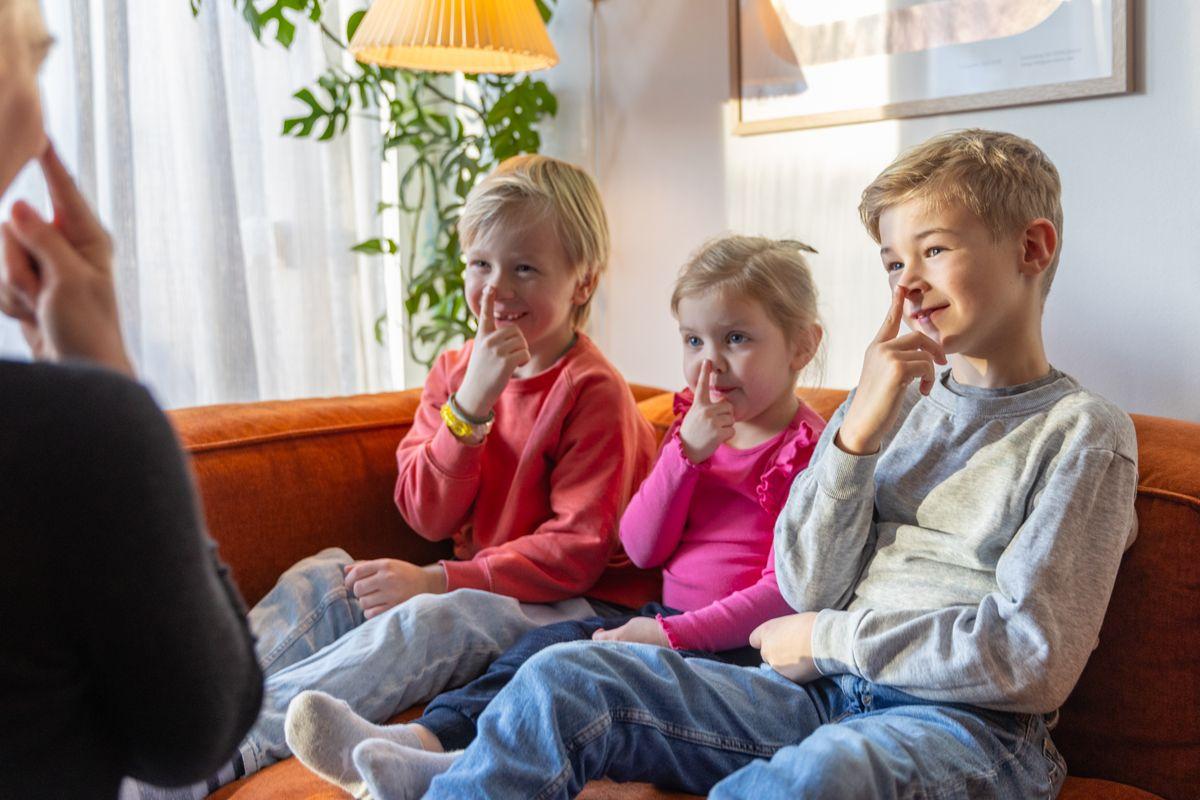Tre barn sitter på en sofa og smiler mens de peker på nesene sine. Sollys strømmer inn i rommet.