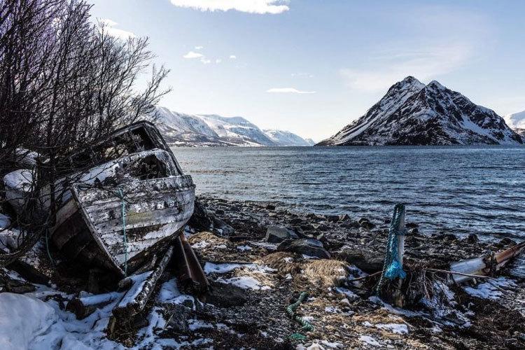 En forlatt båt på en strand med snø, fjell i bakgrunnen og en rolig sjø under en klar himmel.