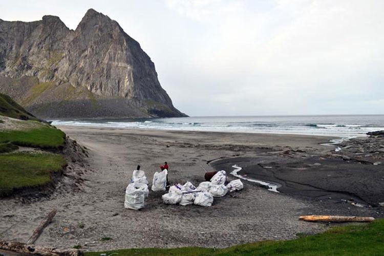 Mennesker i hvite beskyttelsesdrakter rydder søppel på en strand med fjell i bakgrunnen.