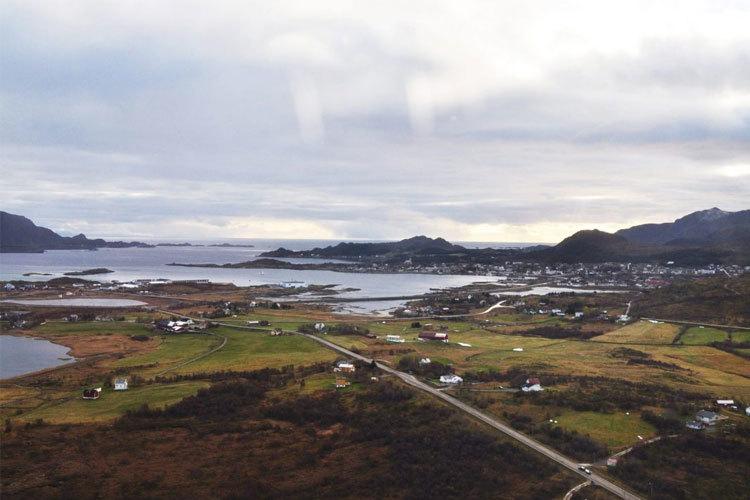 Utsikt over et kystlandskap med hus, grønne enger og fjell i bakgrunnen under en grå himmel.