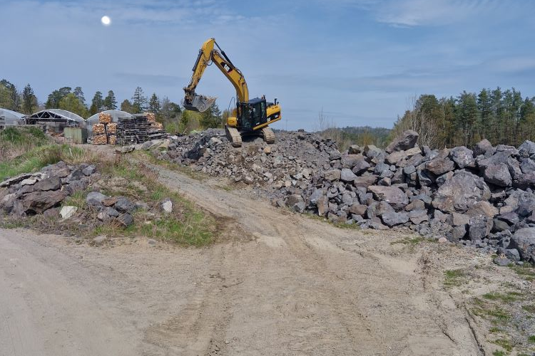 Gravemaskin som arbeider med å flytte stein på en byggeplass med skog i bakgrunnen.