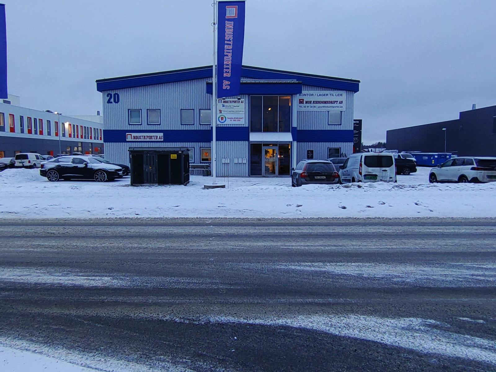 Blå og hvit bygning med flere biler parkert foran, snødekt vei og grå himmel i bakgrunnen.