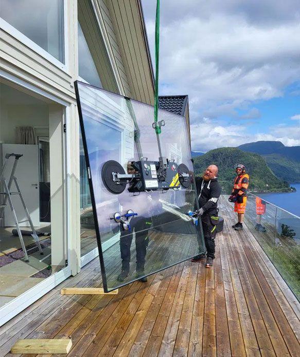 En håndverker heiser et stort glasspanel med kran på en terrasse med utsikt over fjorden.