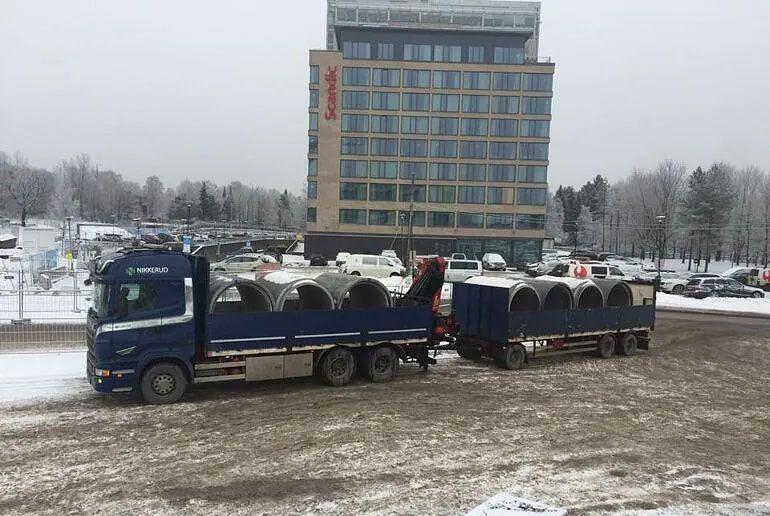 En lastebil med runde betongrør står parkert på en snødekt plass foran et hotell.