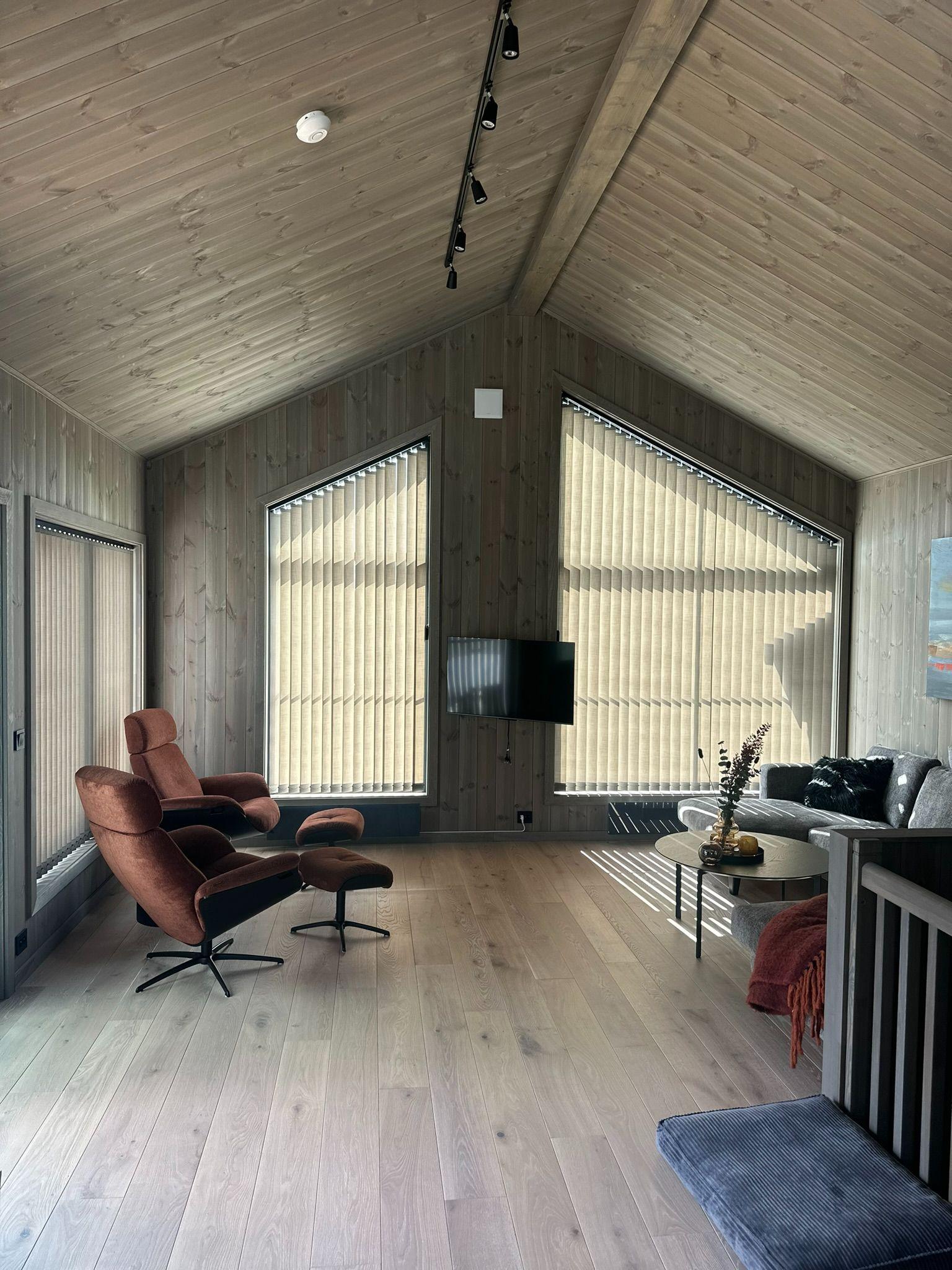 Moderne stue med trevegger, store vinduer, en rød lenestol og en sofa, samt en TV på veggen.