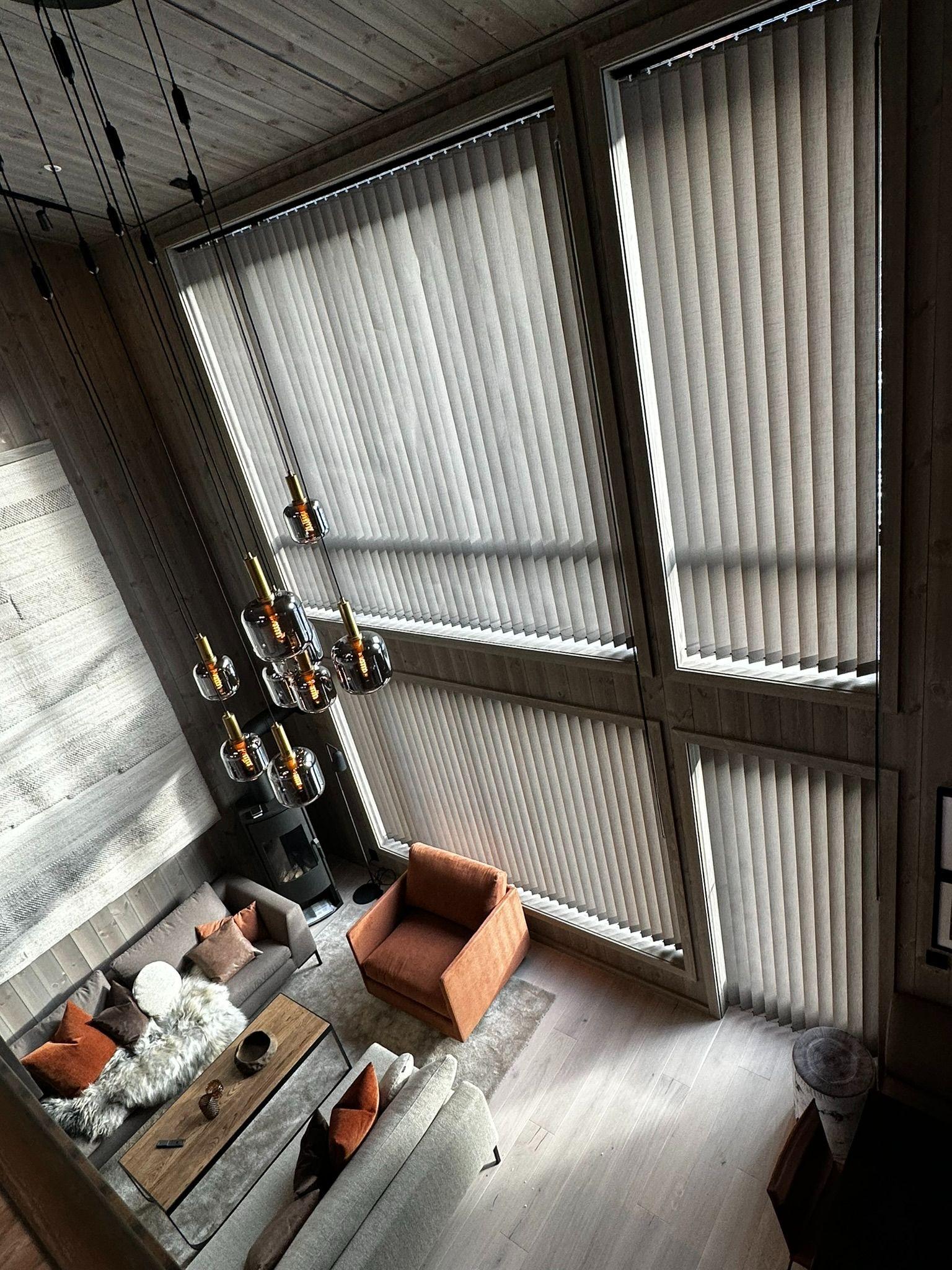 Lys stue med store vinduer, moderne møbler og en stilren lampe hengende fra taket.