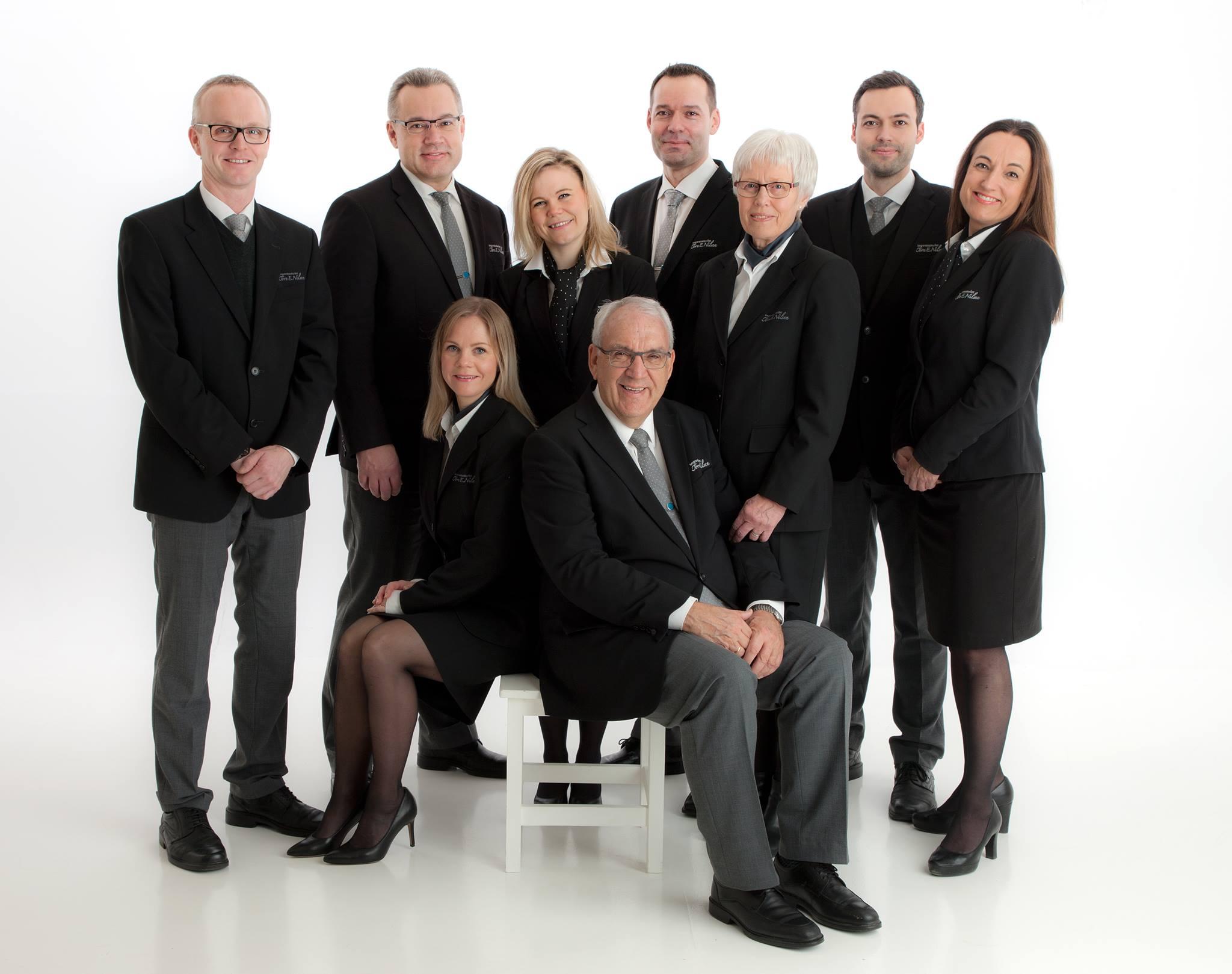 Gruppebilde av ni profesjonelle personer i formelle klær, poserende sammen på en hvit bakgrunn.
