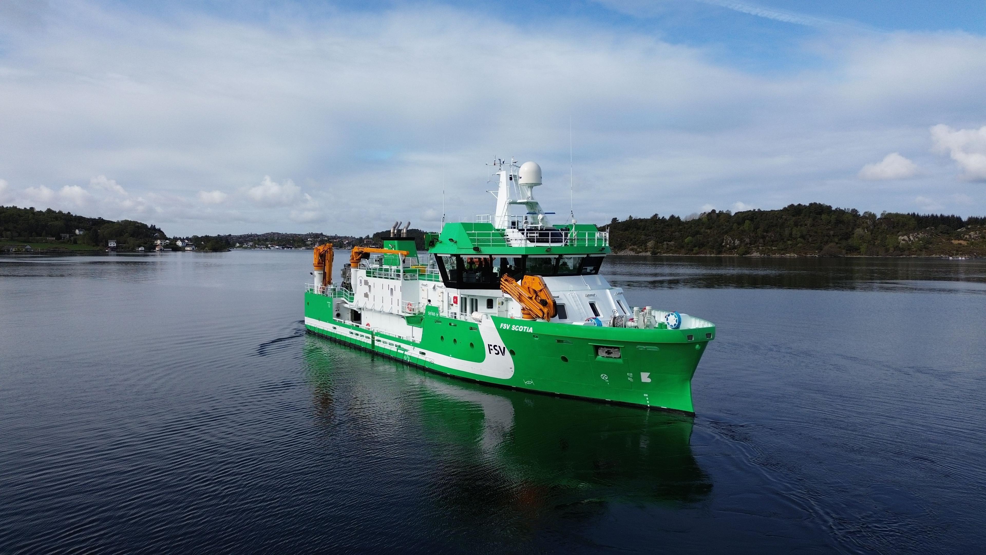 Grønn forskningsskip seiler gjennom rolig vann med kystlinje i bakgrunnen.