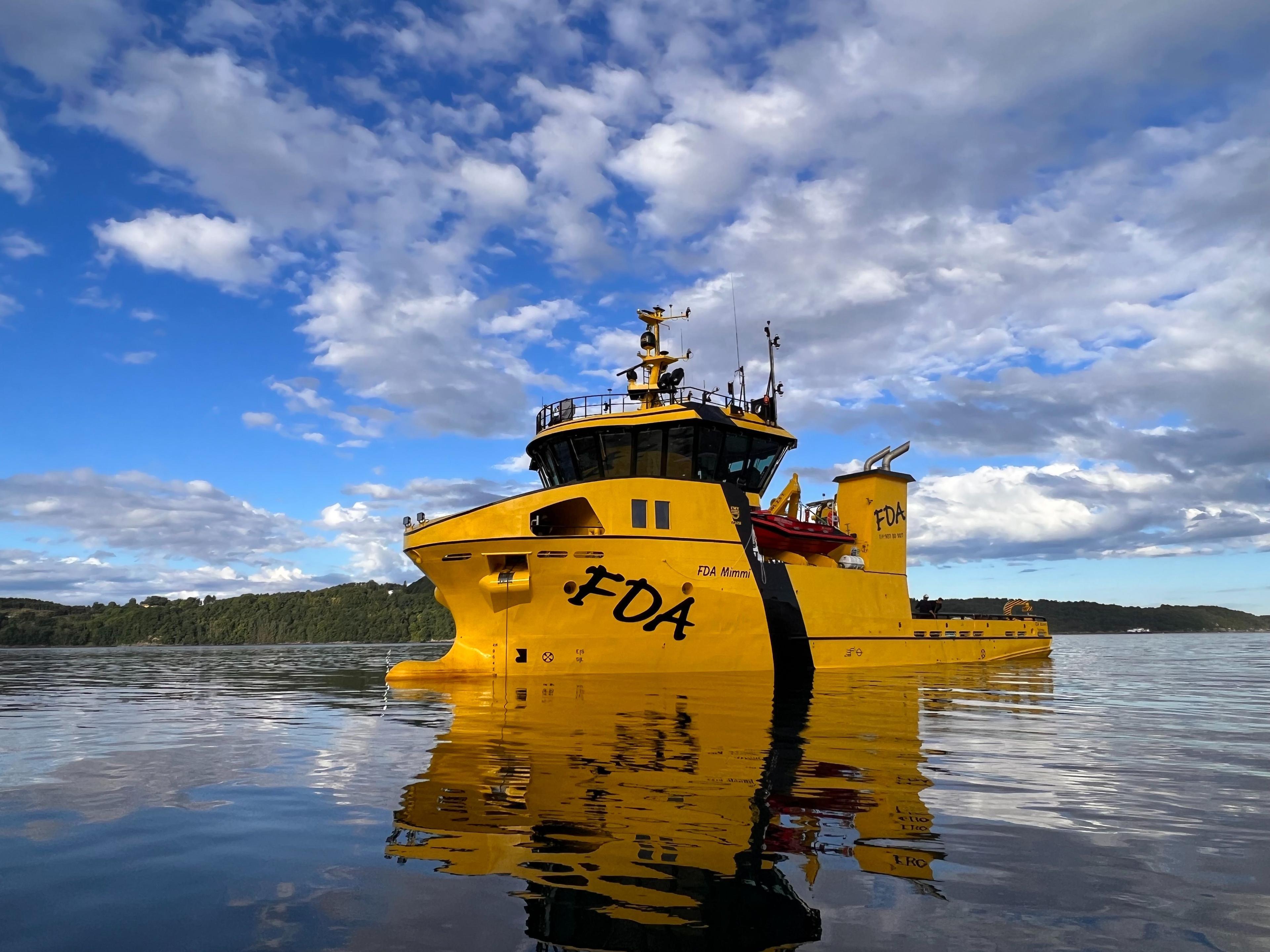 En gul servicebåt med navnet "FDA" ligger stille på vannet under en blå himmel med skyer.