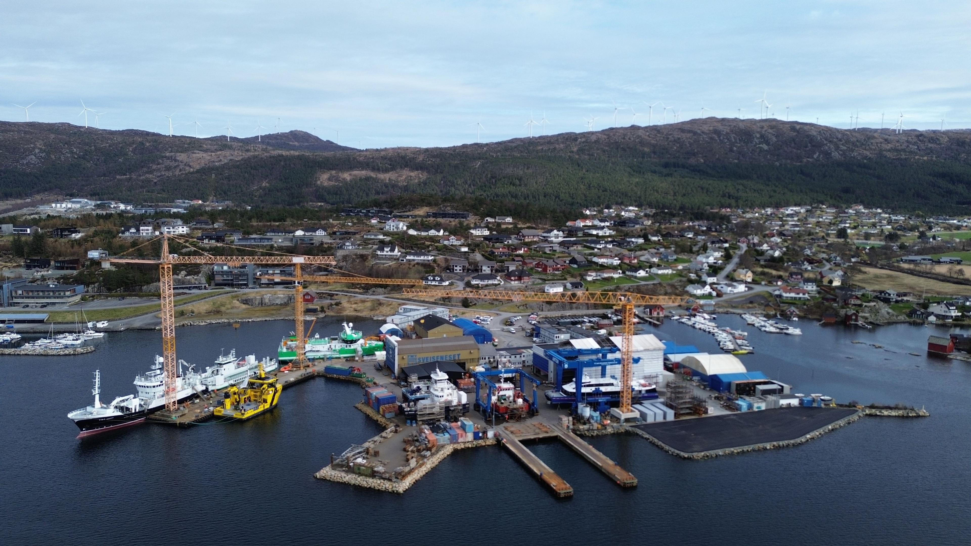 En oversiktsbilde av en havn med flere båter, kraner og bygninger langs vannkanten, omgitt av fjell.