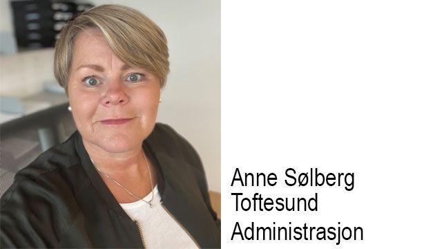 En kvinne med kort, lys hår og en mørk jakke smiler mot kameraet i et kontormiljø.