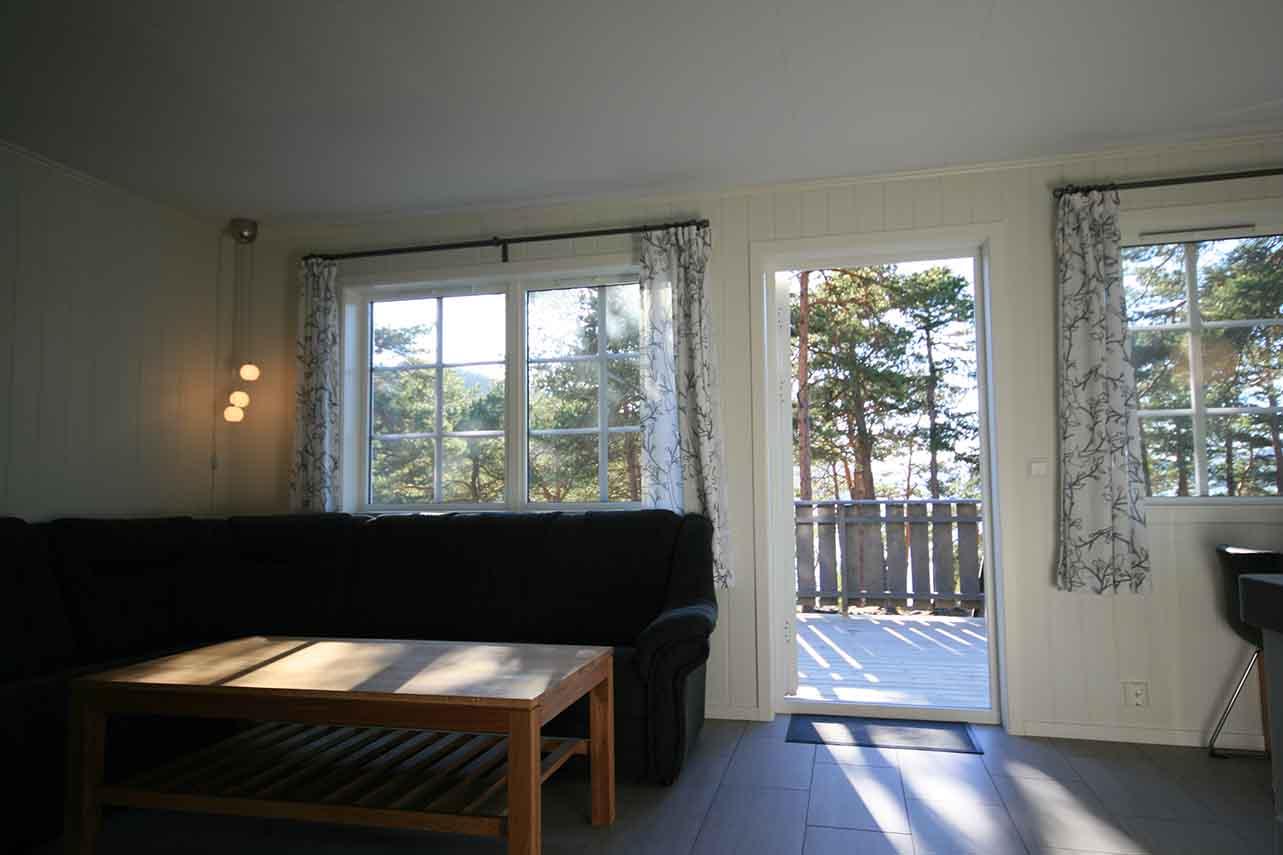 Lys stue med svart sofa, trebord og store vinduer som gir utsikt til en terrasse og trær utenfor.