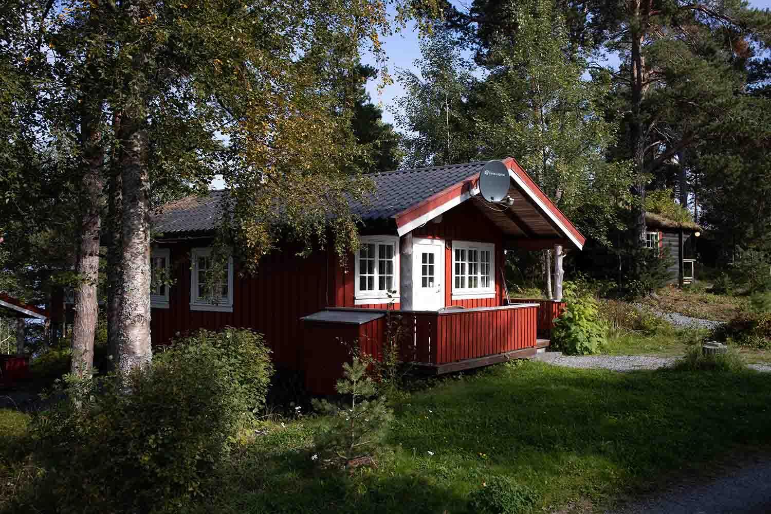 En rød hytte med hvite vinduer, omringet av trær og grønt gress i et naturskjønt område.