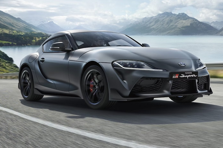 En svart Toyota Supra sportsbil kjører langs en vei med fjell og innsjø i bakgrunnen.