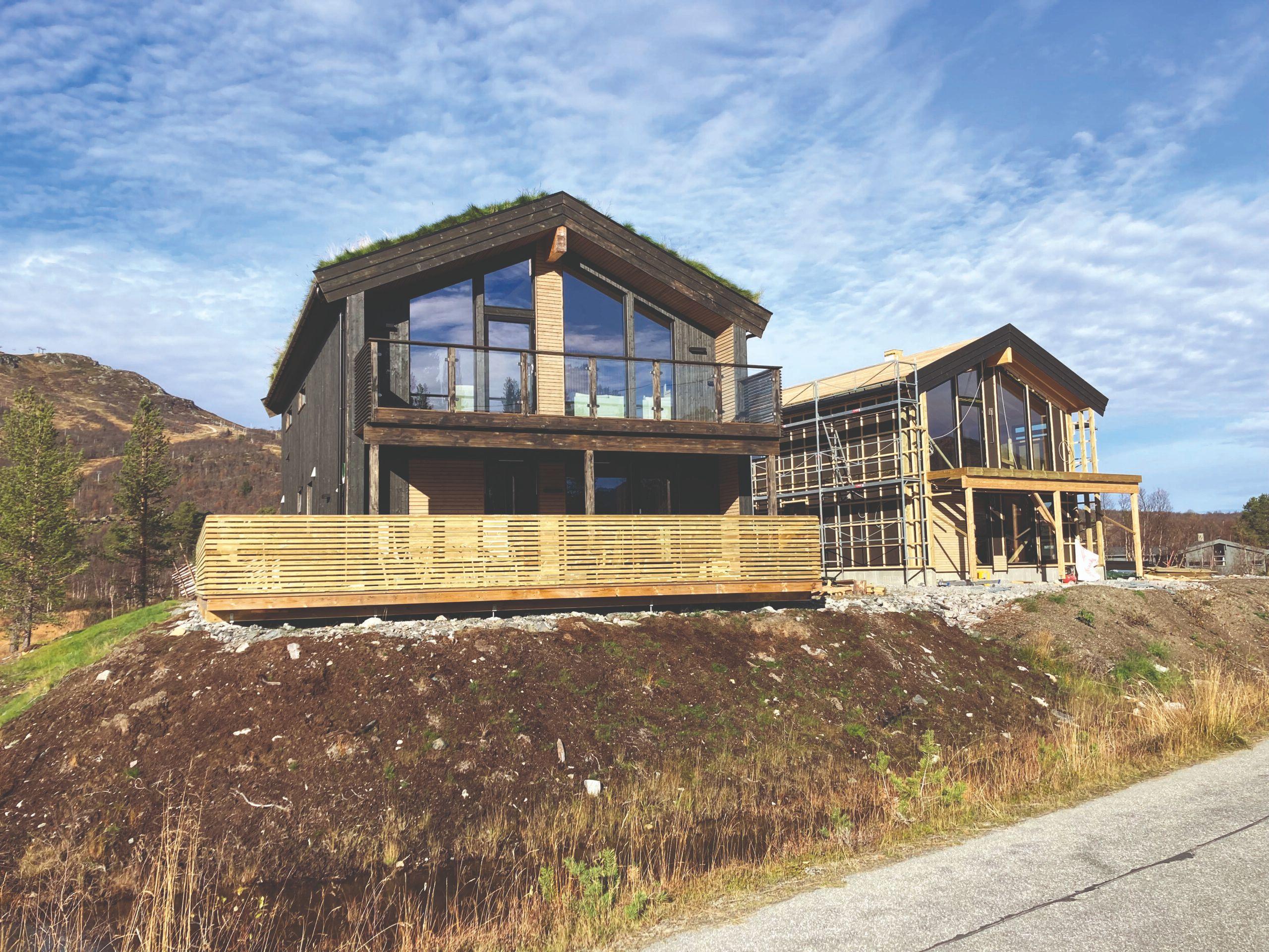 Moderne hytter med store vinduer og terrasse, beliggende i naturskjønne omgivelser under en blå himmel.