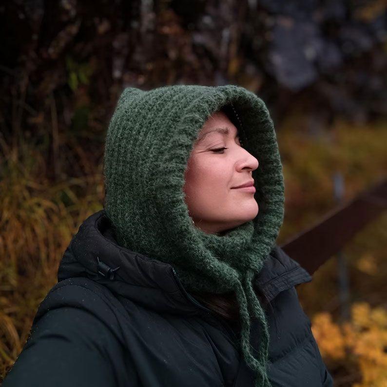 Kvinne med grønn hette og jakke, nyter naturen med lukkede øyne i en høstlig setting.