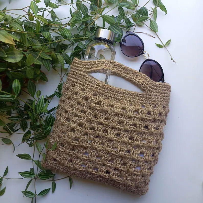 En heklet veske i beige med en vannflaske og solbriller, omgitt av grønne planter.