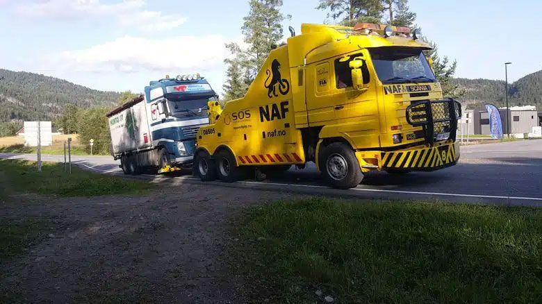 En gul bergingsbil fra NAF hjelper en lastebil som har fått problemer på veien. Grønne fjell i bakgrunnen.