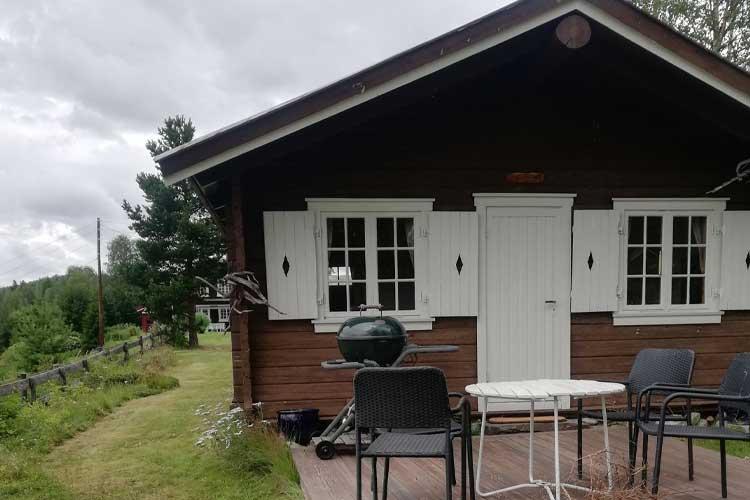 Hytte med hvite vinduer, grill og utemøbler på terrassen, omgitt av grønt landskap og trær.