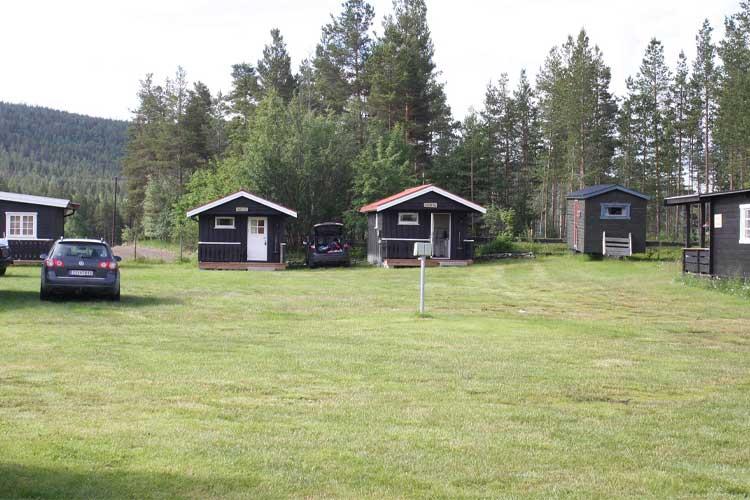 To hytter i en grønn hage med trær i bakgrunnen og en bil parkert mellom dem.