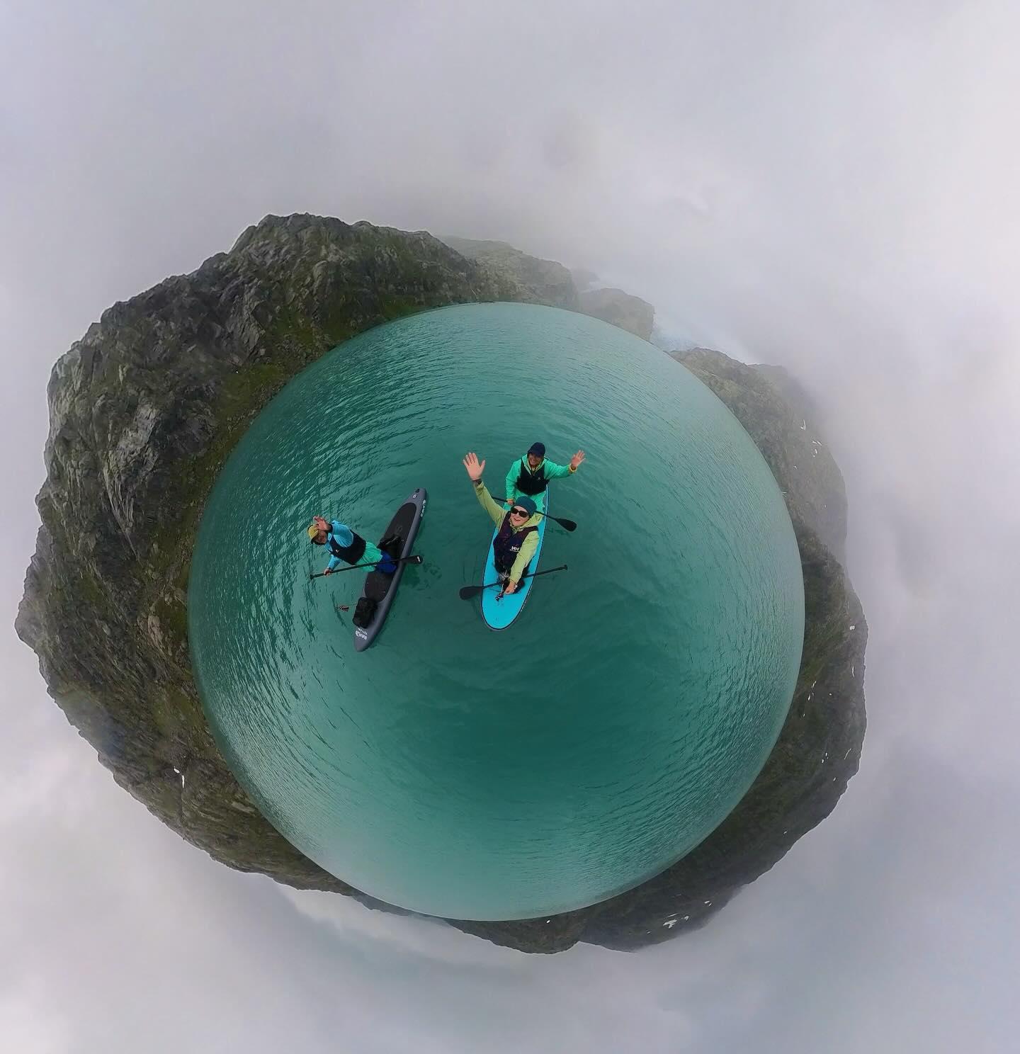 To personer som står på surfebrett i en rund innsjø med klart, turkist vann og fjell i bakgrunnen.