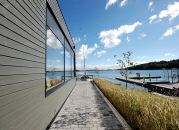 Moderne bygning ved vannet med en steinlagt sti og utsikt over båtplasser og himmel med skyer.