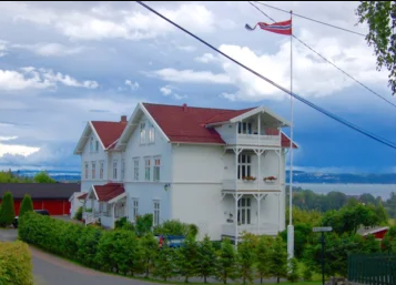 Vakkert hvitt hus med rød tak, flagg på stang, omgitt av grønne busker og utsikt over vannet.